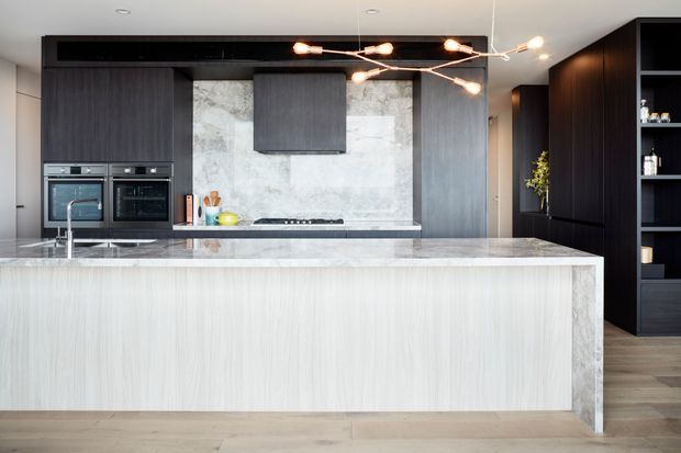 Contemporary open-plan kitchen dominated by a large waterfall-edge island with pale wood-grain cabinetry and a light stone benchtop. The rear wall combines dark grey timber-look cabinetry, integrated appliances, a pale marble-look splashback, and a concealed rangehood. A dark built-in storage wall extends to the right, while warm wood flooring and a sculptural branching pendant light add contrast.