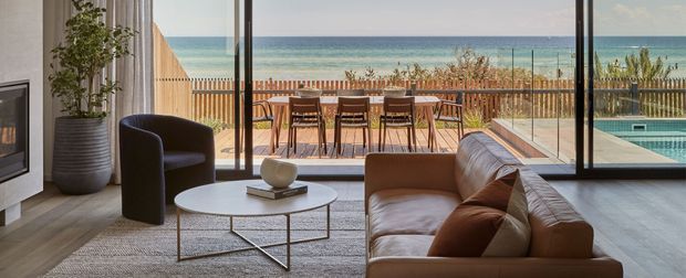Contemporary living room opening onto a beachfront terrace, with a large brown leather sofa, black accent chair, round coffee table, fireplace, potted plant, and full-height glazing. A dining area, pool, deck, beach, and ocean are visible beyond the glass.