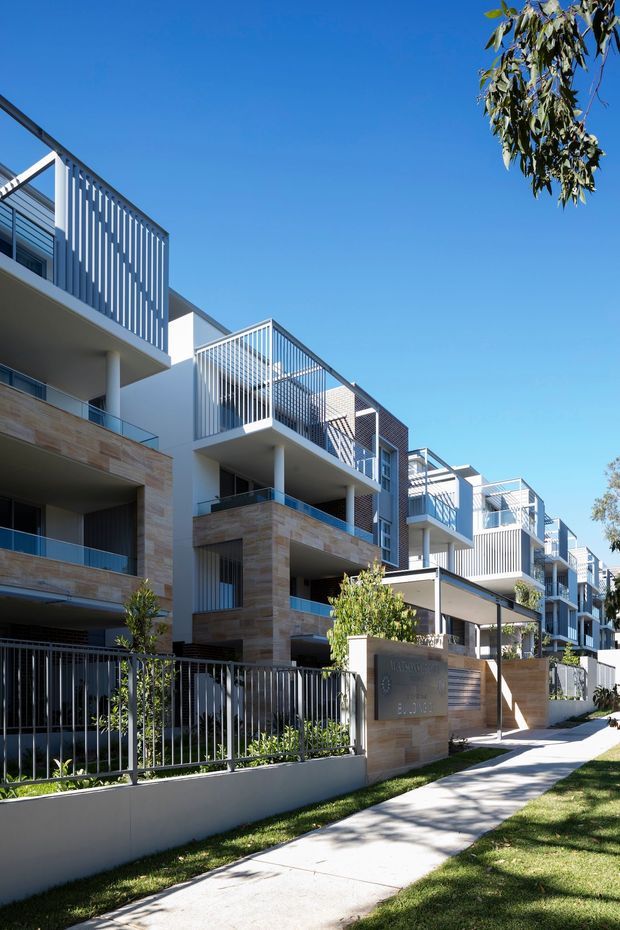 Exterior street-level view of a contemporary multi-storey apartment development. Repeated projecting balconies feature white vertical metal balustrades, pale rendered walls, and warm tan stone-clad sections. A landscaped frontage with fencing, lawn, small trees, and a concrete footpath leads along the building under a clear blue sky.