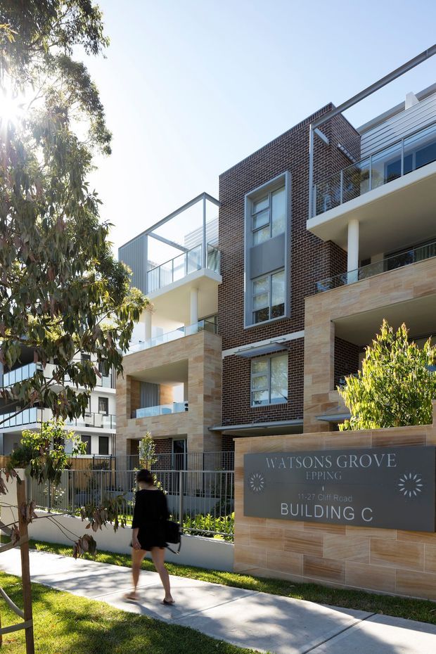 Exterior streetscape view of a contemporary multi-storey apartment development with dark brick facade, light stone-clad sections, glass-railed balconies, projecting terraces, and landscaped pedestrian frontage. A sign identifies the development as Watsons Grove, Epping, Building C.