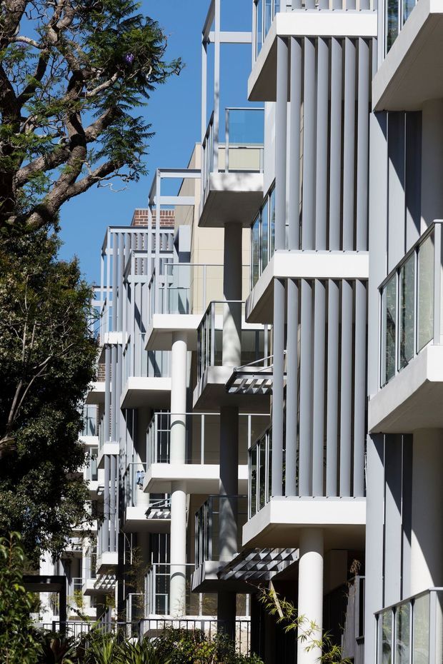 Exterior architectural photograph of a contemporary multi-storey apartment building. The facade is composed of projecting balconies, white vertical fins, extensive glazing with glass balustrades, slender columns, and strong geometric forms. Landscaping and mature trees line the building edge, with a clear blue sky visible.