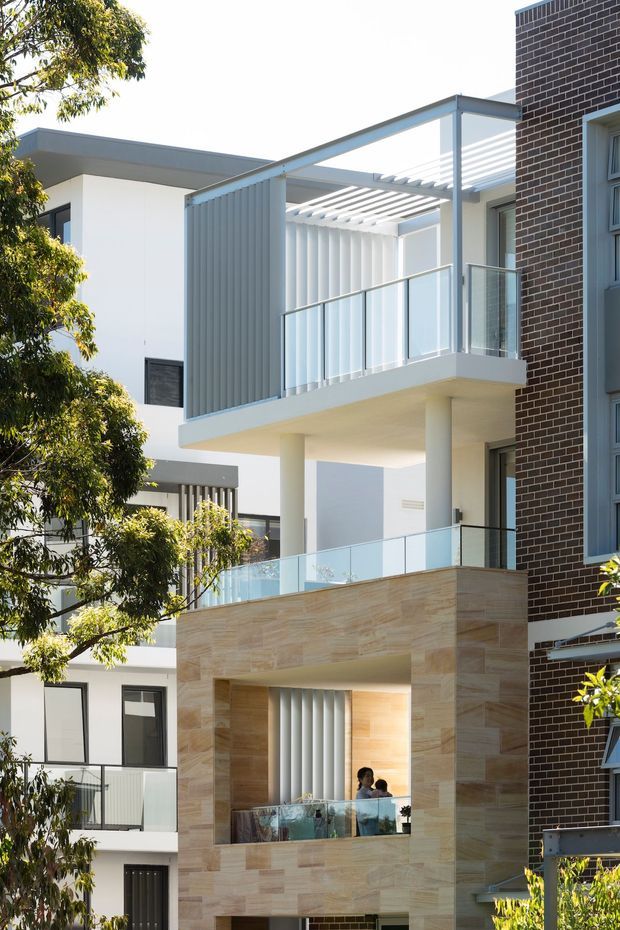 Exterior view of a contemporary multi-storey residential building with projecting balconies, clear glass balustrades, white rendered walls, vertical privacy screens, pergola-style sun shading, and a prominent warm stone-clad central facade. A darker brick-clad neighbouring facade is visible at the right edge, with trees partially framing the scene.