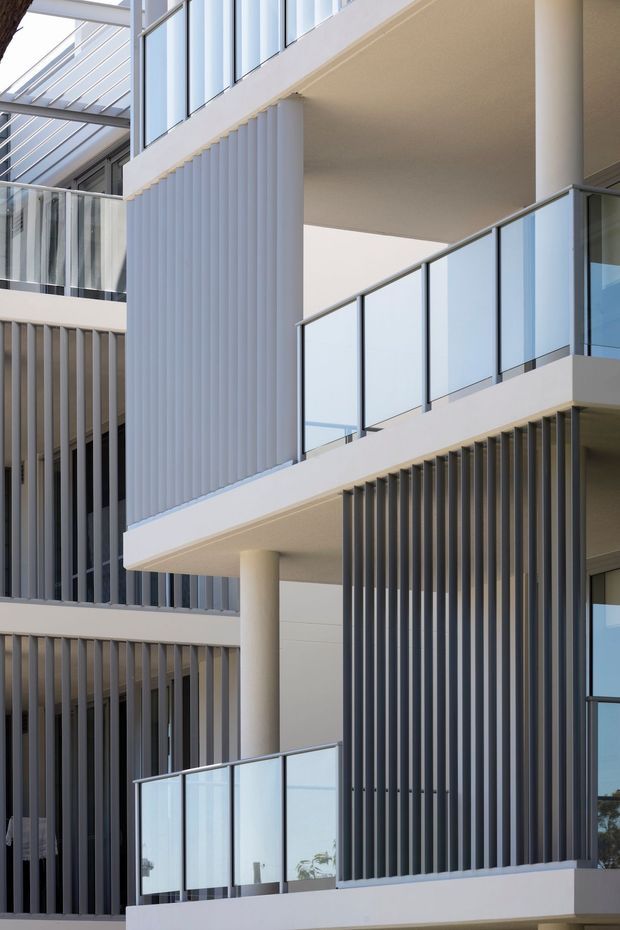 Close-up exterior architectural view of a multi-level contemporary building with stacked balconies, pale concrete or rendered slab edges, transparent glass balustrades, cylindrical columns, and repeated vertical privacy fins in contrasting light and dark tones. The composition emphasizes geometric facade rhythm, layered terraces, and abundant glazing.