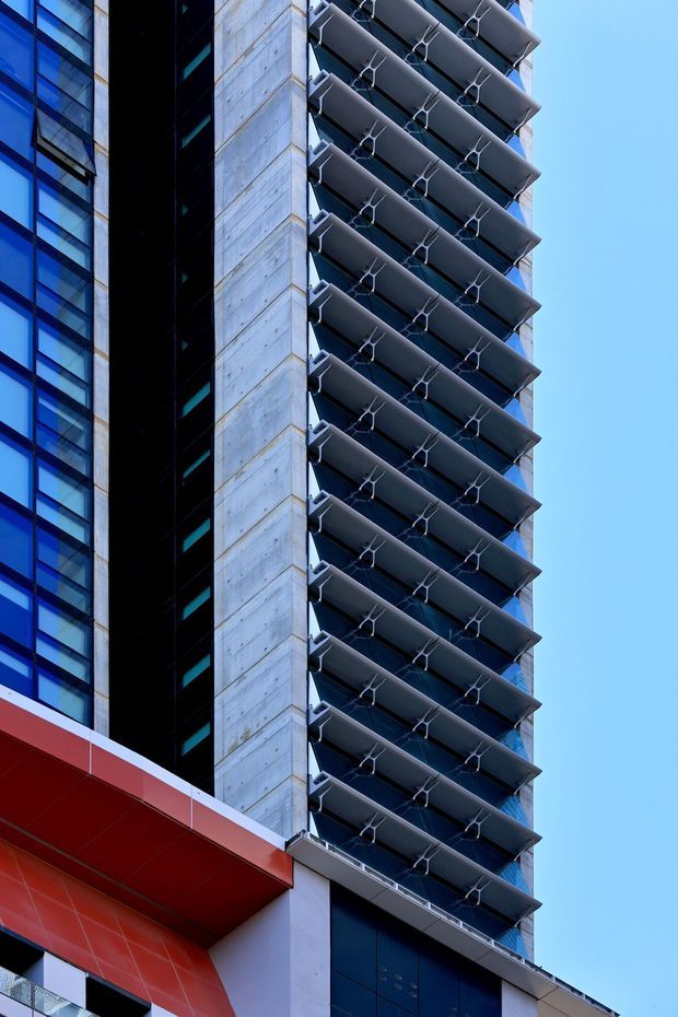 Close-up upward view of a tall contemporary commercial or apartment building facade. The composition highlights repetitive projecting horizontal balcony or sun-shading elements with sculptural metal supports, blue-tinted glazing, a vertical exposed concrete wall, dark recessed sections, and a bold red cantilevered architectural element at the lower left against a clear blue sky.