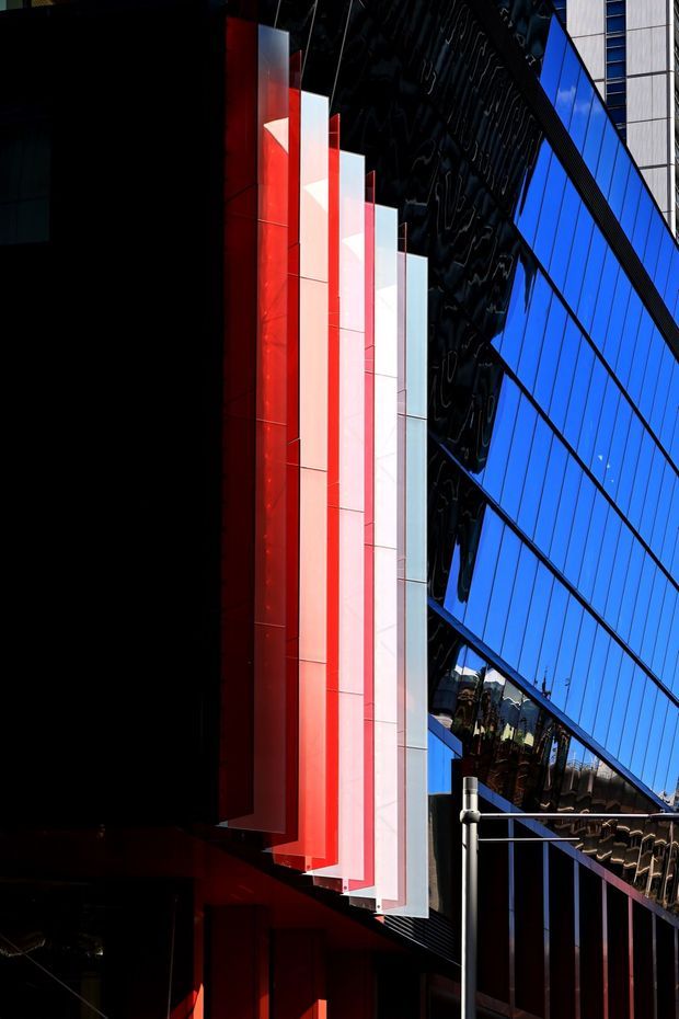 A dramatic close-up view of a contemporary commercial building facade. The composition features reflective blue glass curtain walling, dark glossy cladding, and a striking vertical sequence of projecting red and white fins or panels. Strong sunlight and deep shadows create a bold, high-contrast architectural image.