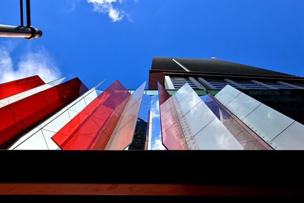 Low-angle exterior view of a striking contemporary multi-storey commercial building. The facade features sharply faceted angular panels in vivid red, white, and reflective silver, contrasted with a darker upper structure and roofline. Bright blue sky and a small portion of an overhead street fixture are visible.