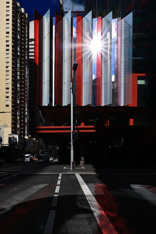 A dramatic urban street-level photograph of a contemporary high-rise building facade. The main structure features projecting angular fins or blades in silver, translucent white, red, and blue, creating a sculptural geometric screen above a dark undercroft. A tall residential tower with rounded balconies stands adjacent on the left. Strong sunlight reflects off the facade, producing high contrast and deep shadows across the street.