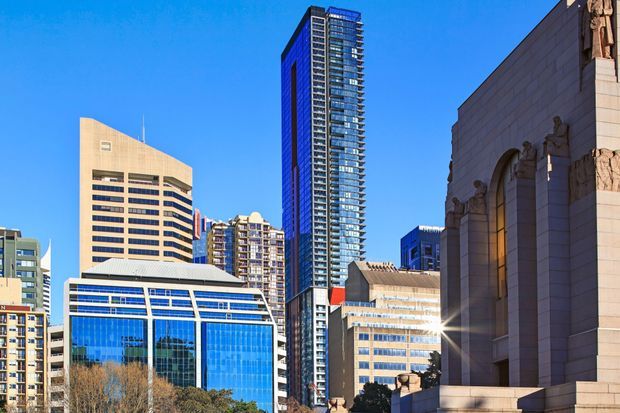 Bright daytime urban architectural view of a dense city centre, featuring a tall slender glass-and-steel residential or office tower, several mid- and high-rise buildings, a modern blue-glazed low-rise building in the foreground, and a large stone-clad monumental facade with sculptural detailing at right.