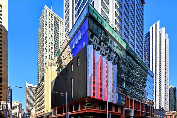 A dramatic urban exterior view of a contemporary high-rise commercial building in a dense city centre. The main structure features a sharply angled glazed facade with reflective curtain walling, dark cladding, vivid blue and red vertical fins, and exposed red structural framing at street level. Numerous surrounding towers and a clear blue sky reinforce the metropolitan setting.