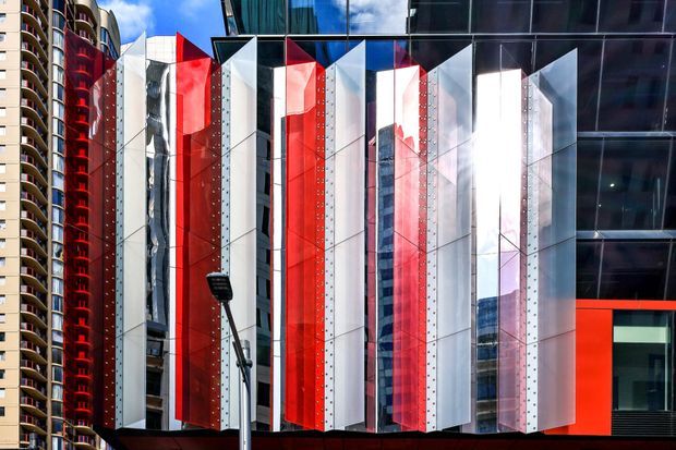 Close-up exterior view of a contemporary urban commercial building facade featuring a dramatic composition of projecting red, white, silver, and reflective glass vertical panels. The geometric fins create strong depth and contrast, with surrounding high-rise buildings reflected in the glazing and visible at the edges.