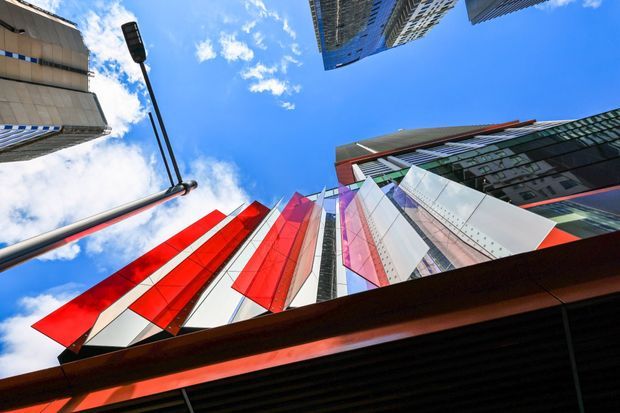 Upward-looking urban architectural photograph showing a dramatic cluster of high-rise buildings around a vivid angular facade. The central building features reflective glass and bold red, white, pink, and grey geometric cladding, with dark projecting structural elements in the foreground. Surrounding towers and street lighting frame a bright blue sky with scattered clouds, creating a dynamic metropolitan composition.
