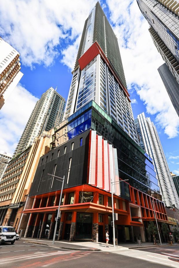 Street-level upward view of a dense urban high-rise building with a dramatic mixed-use facade. The foreground structure combines black cladding, extensive blue-green glazing, bold red-orange framing, vertical white-red panels, and a tall glass-and-metal tower rising above. Surrounding apartment and office towers, streetscape elements, and a bright partly cloudy sky reinforce the metropolitan setting.