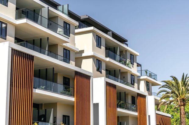 Exterior view of a contemporary multi-storey apartment building with stacked balconies, glass balustrades, white rendered walls, dark roof projections, and prominent vertical timber screening. Palm trees and clear sky are visible around the building.