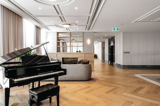 A refined, open-plan interior featuring a glossy black grand piano in the foreground, a curved upholstered sofa, warm timber herringbone flooring, built-in shelving, and a reception or lounge area. The space has high-end contemporary detailing, coffered and recessed ceiling elements, expansive curtained windows, neutral finishes, and a broad circulation zone leading toward a dark marble-accented entry or lift area.