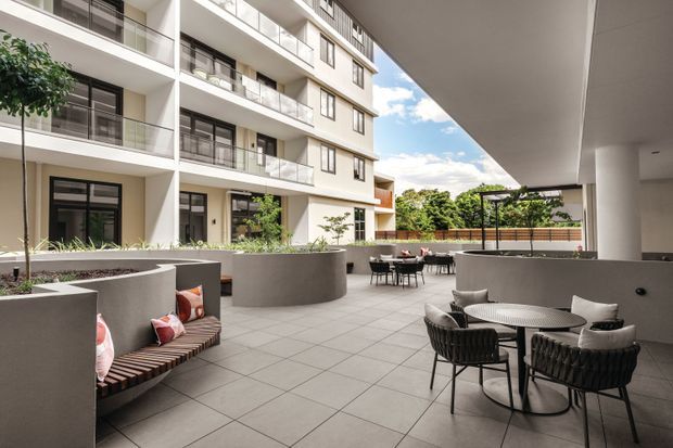 A contemporary apartment courtyard or shared outdoor terrace with multi-storey residential buildings, recessed balconies, glass balustrades, and dark-framed glazing. The paved space includes curved raised planters, integrated timber seating, café-style tables and chairs, and a pergola-covered dining area. The palette is predominantly light grey, white, and charcoal with greenery providing contrast.