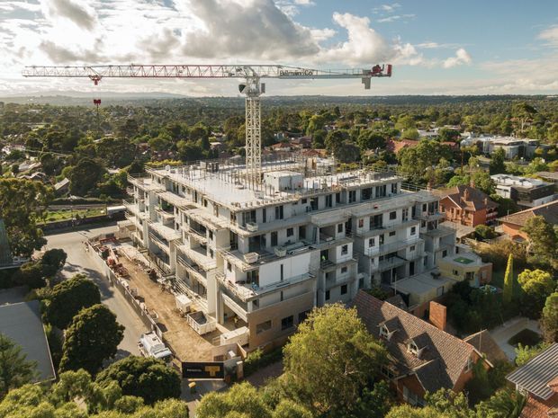 Aerial view of a multi-storey residential or mixed-use building under construction, with exposed concrete floor slabs, structural columns, incomplete facade elements, and a tower crane. The development is situated in a leafy suburban neighbourhood with surrounding houses and streets under a partly cloudy sky.