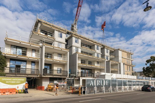 Street-level exterior photograph of a multi-storey apartment building under construction, with exposed concrete structure, balconies, window openings, construction hoarding, scaffolding, and a tower crane. The building has a contemporary flat-roofed form with repetitive balconies and light-coloured facade elements.