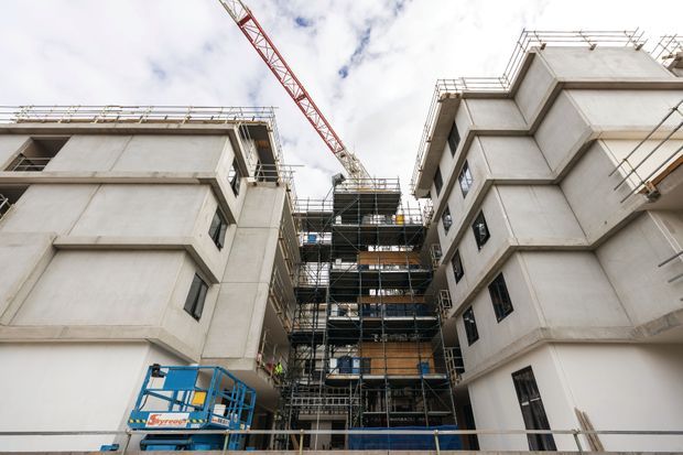 Exterior construction-site view looking between several multi-storey concrete building volumes. The structures are unfinished, with exposed concrete facades, scaffolding, temporary edge protection, open window openings, construction equipment, and a tower crane under a cloudy sky.