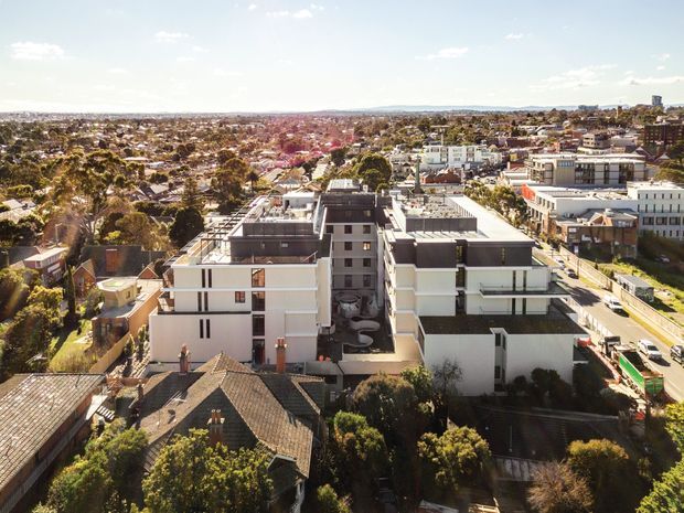 Aerial view across a dense suburban-urban neighbourhood, with a modern multi-storey white apartment development in the foreground surrounded by established low-rise homes, mature trees, and commercial or institutional buildings. The scene is brightly lit under a clear sky and shows a predominantly flat urban setting.