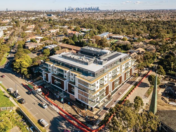 Aerial view of a contemporary multi-storey apartment building under construction, with white and dark grey facade elements, projecting balconies, rooftop terraces and extensive glazing. The building sits within an established suburban neighbourhood, with a dense city skyline visible in the distance and construction fencing around the site.