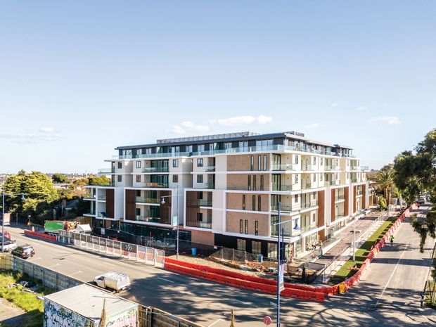 Exterior aerial-oblique view of a contemporary multi-storey apartment building under construction on a corner urban site. The building has a stepped, flat roofline, extensive balconies, large glazed openings, white rendered sections, and warm brown brick facade panels. Construction fencing, temporary barriers, and unfinished landscaping surround the site, with established suburban streets and trees nearby.
