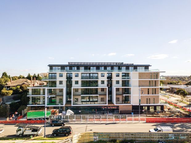 Exterior view of a contemporary multi-storey apartment building under construction, featuring a long flat roof, white rendered and brick facade sections, extensive glazed balconies, dark window frames, and ground-level commercial or communal spaces. Construction fencing and works are visible in the foreground, with suburban residential buildings surrounding the site.