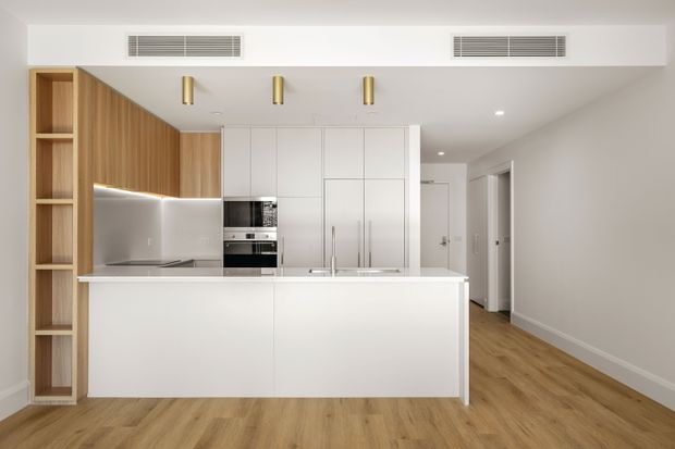 Bright, contemporary open-plan kitchen with a large white island, integrated white cabinetry, stainless-steel appliances, a pale reflective splashback, and warm timber-look flooring. Vertical timber shelving and overhead timber cabinetry add warmth, while a hallway extends beyond the kitchen.