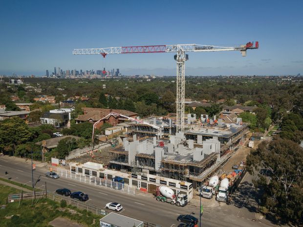 Aerial view of a multi-storey building under construction, with a prominent tower crane, concrete structural frame, scaffolding, construction vehicles, and site fencing. The project is set in an established suburban area with a dense city skyline visible in the distance.