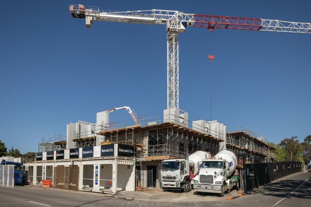 Photograph of an active multi-storey building construction site with a tower crane, concrete mixer trucks, scaffolding, temporary site offices and partially completed concrete structural framing under a clear blue sky.