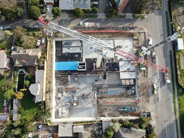 Aerial top-down view of an active multi-storey building construction site in a suburban neighbourhood. The site includes a large concrete slab and partially constructed building, tower cranes, construction vehicles, scaffolding, temporary materials, and surrounding residential properties and streets.