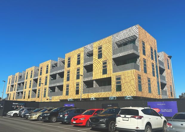 Exterior view of a multi-storey contemporary apartment building under construction or recently completed, with a striking yellow patterned brick facade, dark recessed balconies, and tall narrow windows. The building presents a repeated modular form with multiple levels of apartments and is photographed from a street-level parking area. Parked vehicles and construction hoarding occupy the foreground beneath a clear blue sky.