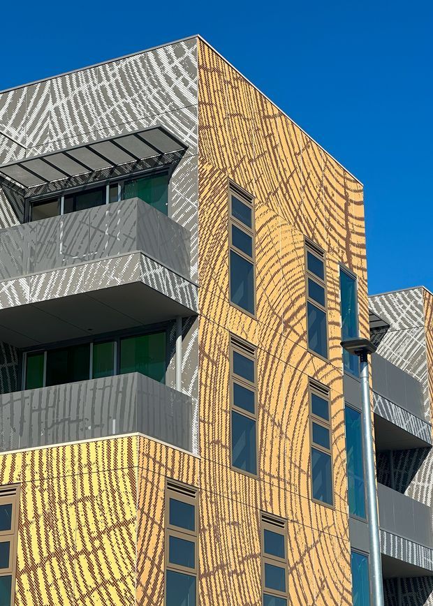 Close-up exterior view of a contemporary multi-storey apartment building with striking patterned yellow and grey facade cladding. Narrow vertical windows, projecting balconies with grey metal balustrades, a glazed canopy, and prominent exposed services or downpipes are visible against a clear blue sky.