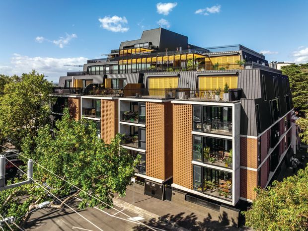 Exterior view of a contemporary multi-storey apartment building with stepped terraces, dark metal cladding, exposed warm brick screens, extensive glazing, balconies, rooftop planting, and a flat-roofed upper form. The building is set in an urban streetscape with mature trees and visible utility wires.