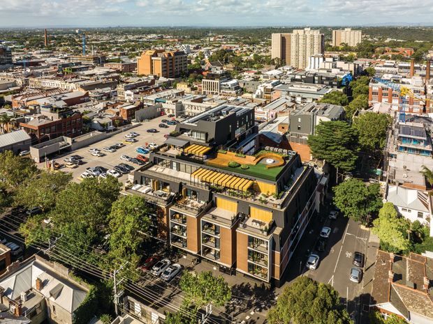 Aerial view of a dense urban neighbourhood featuring a contemporary multi-storey apartment or commercial building in the foreground, with rooftop terraces, yellow awnings, planted roof areas, and surrounding streets, parking lots, low-rise buildings, and taller apartment towers.