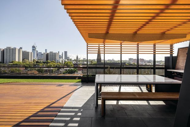 A contemporary rooftop terrace or balcony with a large timber pergola, slatted screening, timber decking, dark tiled paving, and an outdoor dining table with bench seating. The open edge overlooks a dense urban skyline, while strong sunlight creates patterned shadows across the terrace.
