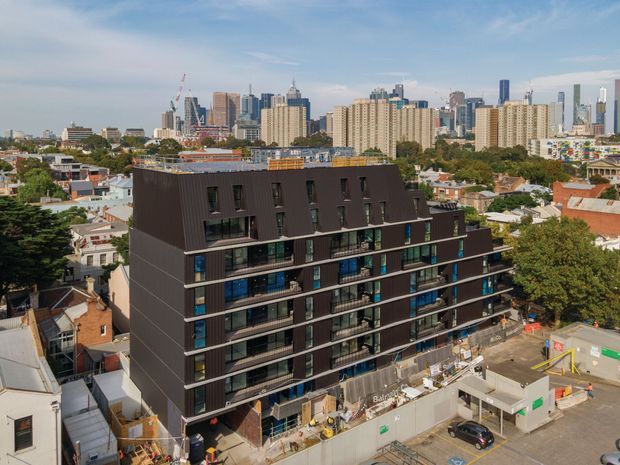 Aerial view of a contemporary multi-storey apartment building under construction in a dense urban neighbourhood. The building has a dark charcoal facade, regularly spaced balconies and glazing, and a steeply sloped dark roof-like upper section. A high-rise city skyline and surrounding low-rise residential buildings are visible in the background, with construction fencing and equipment around the site.