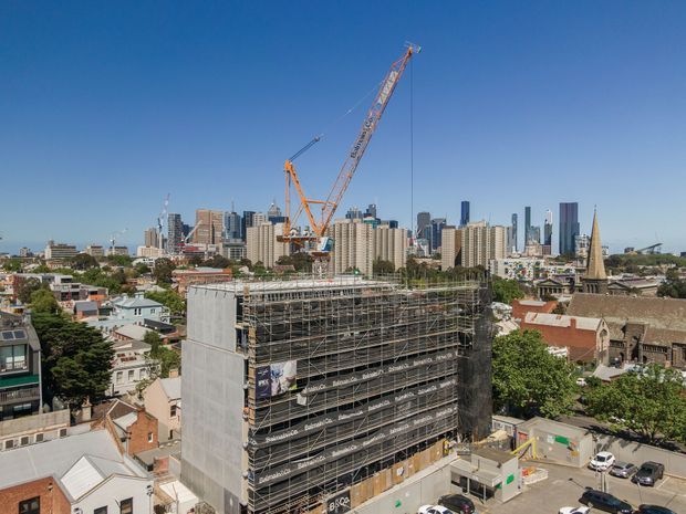 A daytime elevated view of a dense urban construction site, with a multi-storey building wrapped in scaffolding and safety mesh in the foreground. Tower cranes rise above the site, while a city skyline, low-rise residential and commercial buildings, and a prominent church spire extend across the background under a clear blue sky.
