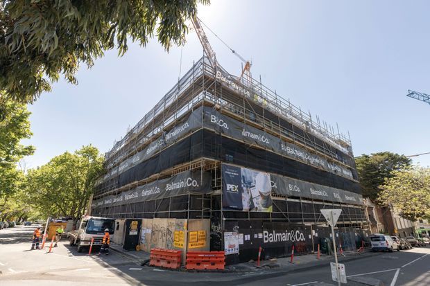 A multi-storey urban building under construction, enclosed in extensive scaffolding and black construction wrap marked with contractor branding. Cranes, temporary barriers, construction signage, workers, and partially visible surrounding street trees and buildings are present.