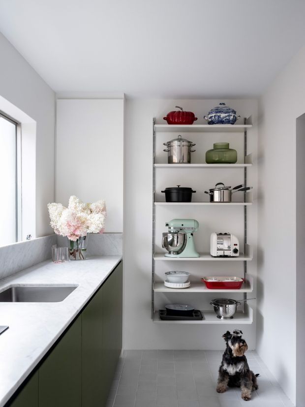Bright, compact kitchen or pantry area with a white marble-look benchtop, integrated sink, dark green lower cabinetry, and open wall-mounted shelving displaying cookware and small appliances. A large window provides natural light, while a small dog sits on the grey tiled floor.