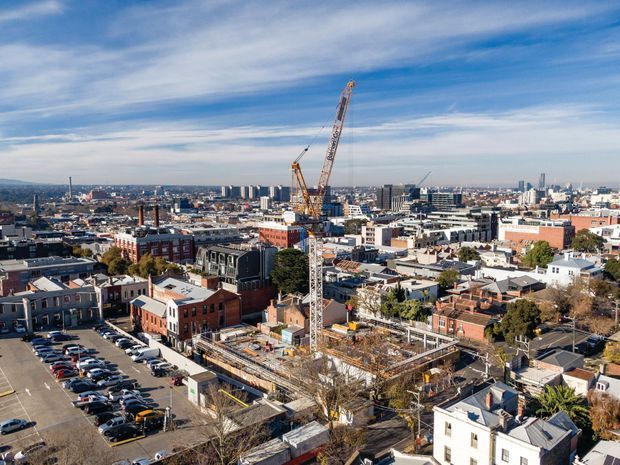 Wide elevated view over a dense urban district with a large active building construction site in the foreground, including tower cranes, exposed concrete framing, and partially completed structures. Low-rise brick and painted buildings, parking areas, streets, and taller apartment or commercial buildings extend across the cityscape under a bright blue sky.