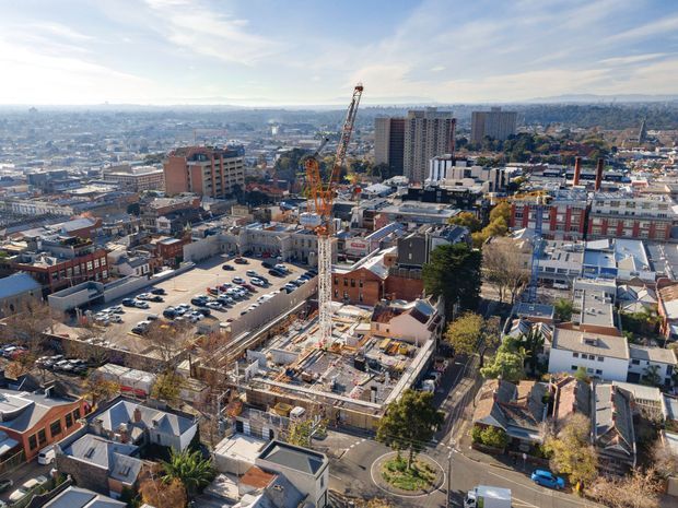 A high-angle aerial view over a dense urban area, showing a mix of low-rise commercial and residential buildings, several taller apartment or office buildings, active construction with tower cranes in the foreground, streets, parking areas, and distant hills under a bright sky.