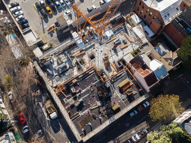 Aerial view of a dense urban construction site showing a multi-storey concrete building under construction, with exposed slabs, reinforcing steel, formwork, scaffolding, construction workers, and a prominent orange tower crane. Surrounding streets, parked cars, and neighbouring buildings are visible.