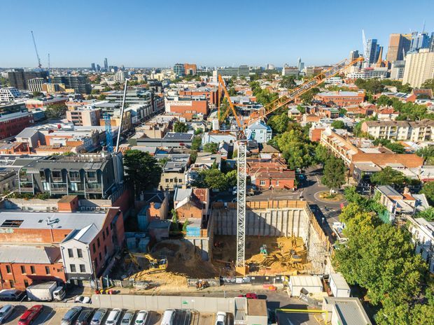 Elevated panoramic view over a dense urban neighbourhood with extensive construction activity, including tower cranes, an excavation site, mid-rise commercial and residential buildings, and a prominent high-rise skyline in the background. The scene is a bright, clear daytime cityscape.