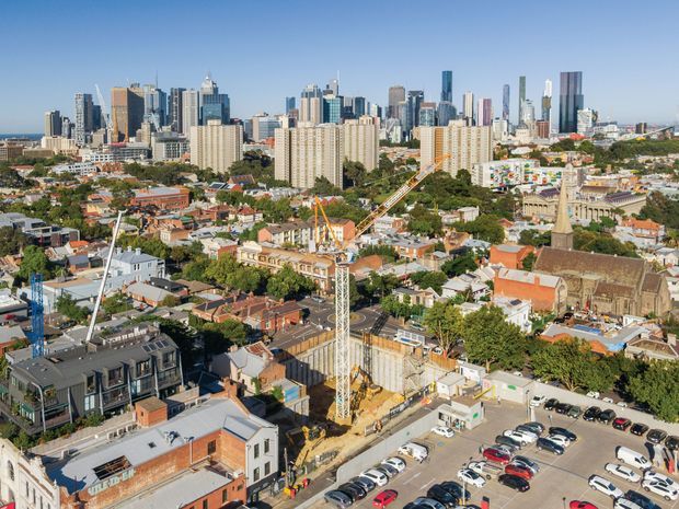 Wide elevated cityscape showing a dense urban skyline of modern high-rise towers behind a mixed residential and commercial neighbourhood. Older low-rise buildings, a prominent historic church, construction activity with a tower crane, streets, and a large parking area are visible in the foreground.