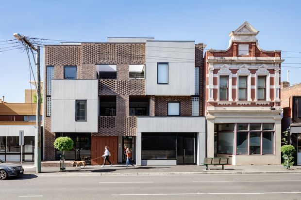Street-facing architectural view of a contemporary multi-storey infill building adjoining a restored ornate Victorian-era commercial facade. The new building combines exposed reddish-brown brick latticework, smooth light-grey cladding, large dark-framed windows and recessed balconies, while the neighbouring heritage facade features decorative mouldings, columns and a parapet. Urban streetscape with overhead utility wires and a broad paved road.