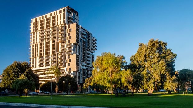 Exterior photograph of a tall contemporary apartment building set beside a broad landscaped urban park. The multi-storey tower has a rectilinear form, pale masonry or concrete facade panels, extensive balconies, and repetitive vertical window bands. Mature trees, lawn, street lighting, and a clear blue sky surround the building.