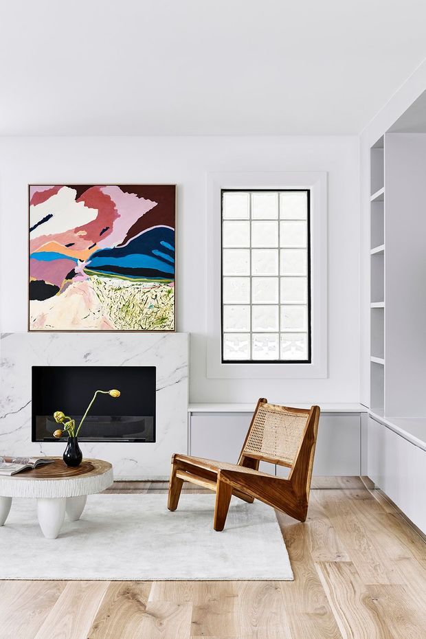 Bright, refined living room with white walls and built-in shelving, a marble fireplace or media surround, pale timber flooring, and a large abstract artwork. A sculptural round coffee table, black television, and woven wooden lounge chair create a clean contemporary setting with mid-century influences.