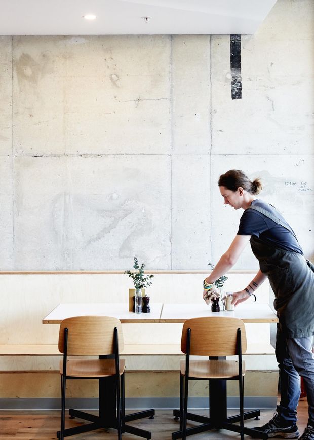 Bright minimalist café or casual dining space with a pale concrete feature wall, built-in light timber banquette, white rectangular communal table, and two matching timber-and-black-metal chairs. A staff member is arranging small table plants and condiments. The room has a clean, utilitarian fit-out with exposed concrete and warm timber details.