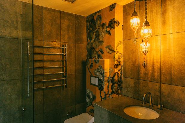A moody, richly detailed bathroom featuring dark large-format wall tiles, a mosaic-tiled structural column, a wall-mounted chrome heated towel rail, a floating dark vanity with an integrated oval basin, and ornate amber pendant lights. The warm lighting and decorative mosaic create a luxurious, dramatic atmosphere.
