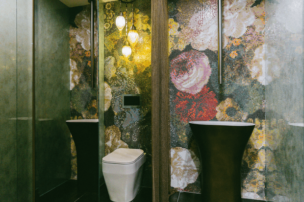 A compact, dramatic bathroom featuring a white toilet and two black pedestal basins or vessel sinks. The walls are clad in dark reflective surfaces and a highly decorative mosaic feature wall with large abstract floral and geometric imagery. Pendant lights hang in front of the mosaic, creating a luxurious, theatrical atmosphere.